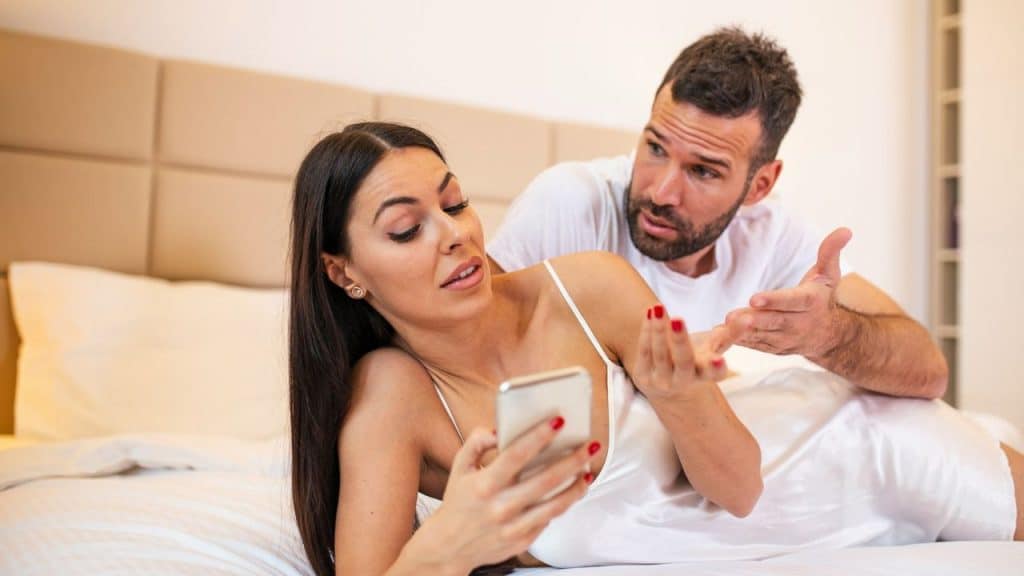 A woman on a bed looks annoyed at a man who gestures while looking over her shoulder.