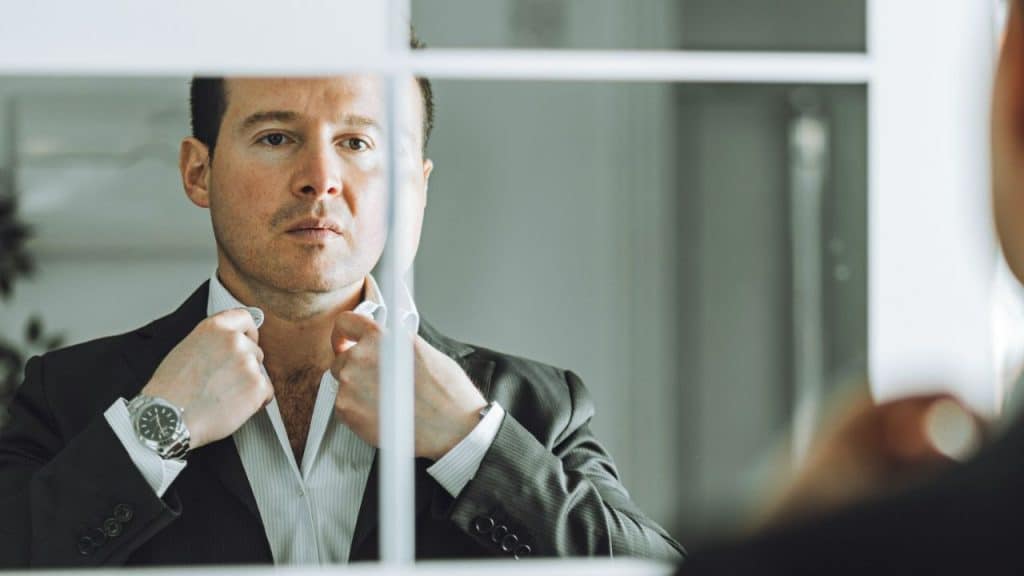Man in a dark suit looking into a mirror and adjusting his shirt collar.