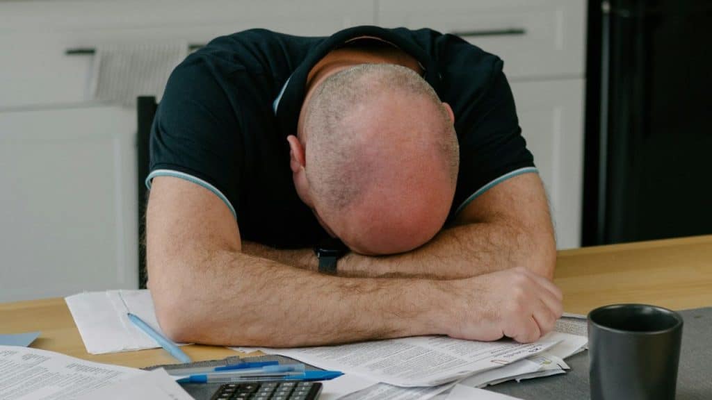 A bald man rests his head on his arms on a table, surrounded by documents and a calculator.