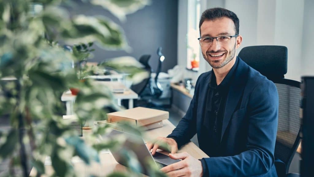 Smiling man in a blue blazer and glasses working at a desk in an office.