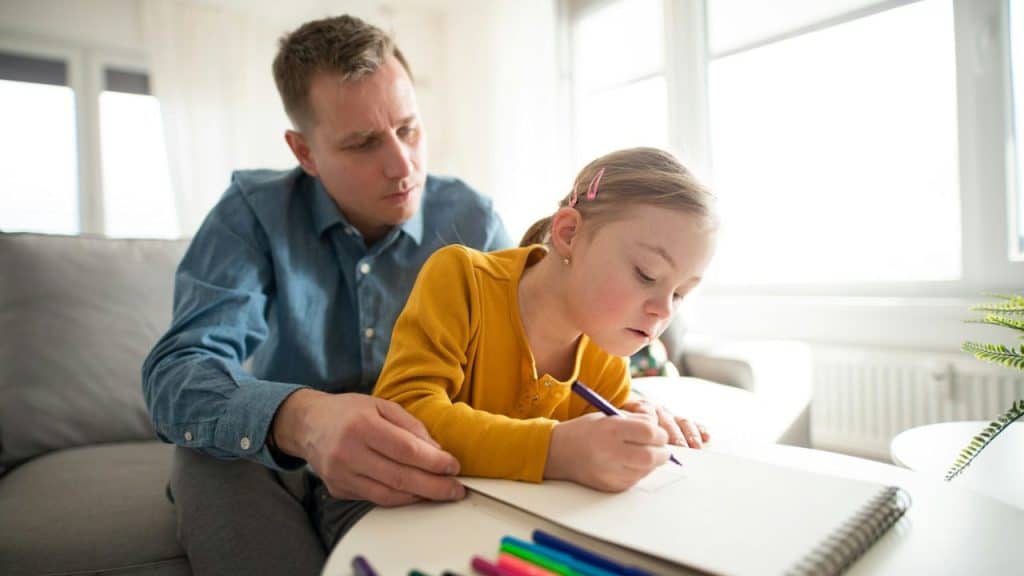 Father and young daughter drawing together on a couch in a bright room.
