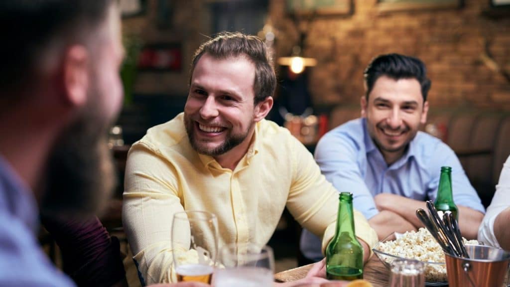 Three bearded men smiling and socializing around a table with drinks and popcorn.