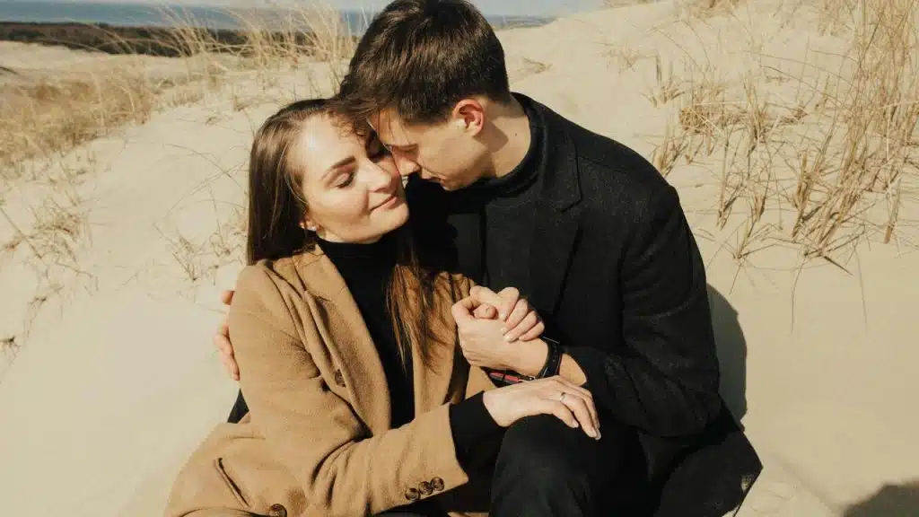 A couple sitting close together on sandy dunes while holding hands.