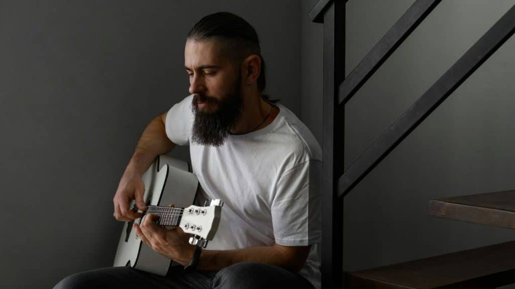 Bearded man in a white shirt playing a white acoustic guitar near stairs.