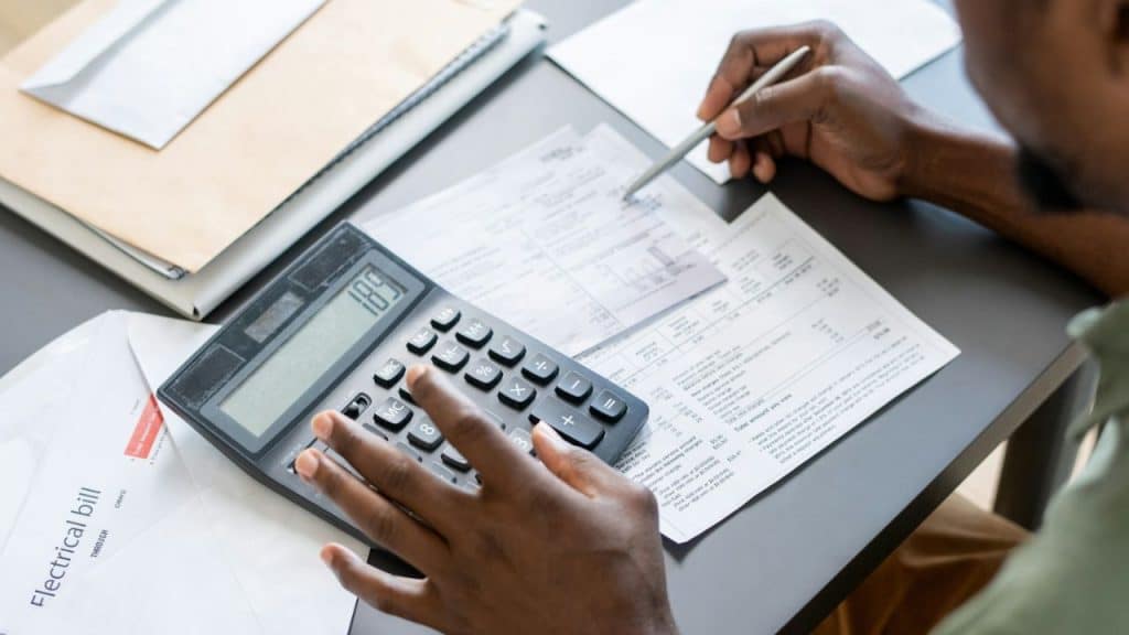 Person using a calculator and pointing a pen at papers on a gray desk.