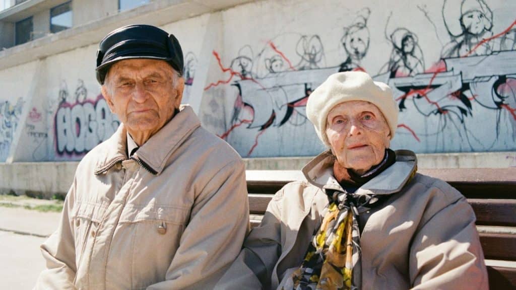 Elderly couple in coats and hats sitting on a bench in front of a graffiti wall.