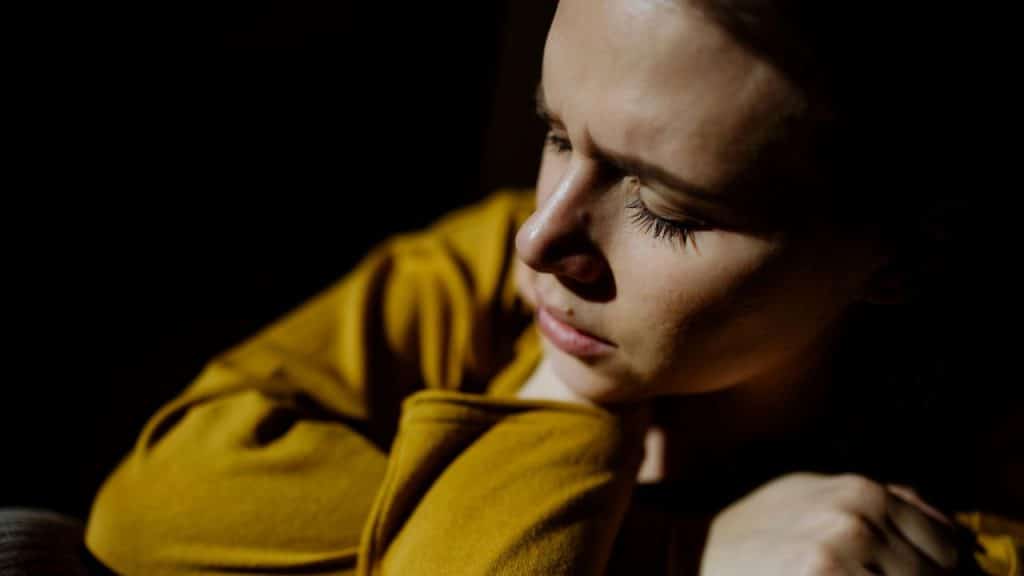 Woman in a mustard-yellow top looking down, with bright light highlighting her face.