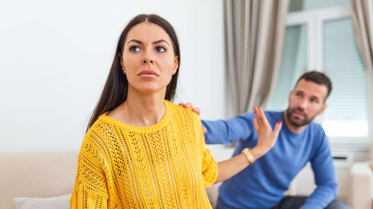 Woman in yellow looking away coldly while a man behind her gestures in protest.