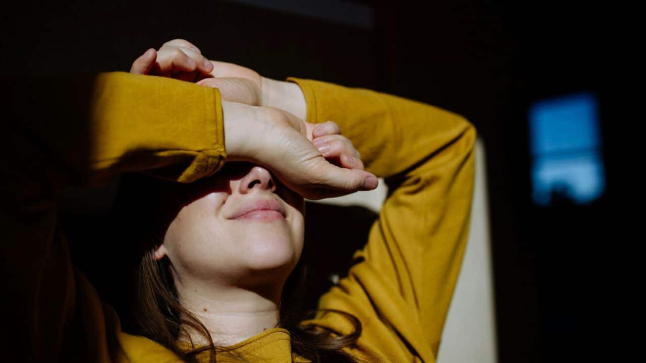 Woman in a yellow shirt covering her eyes with her hands, in bright light against a dark background.
