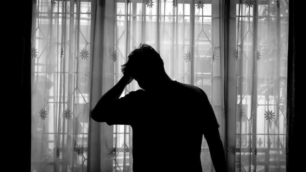 A silhouette of a person standing by a window with sheer curtains, holding their head.