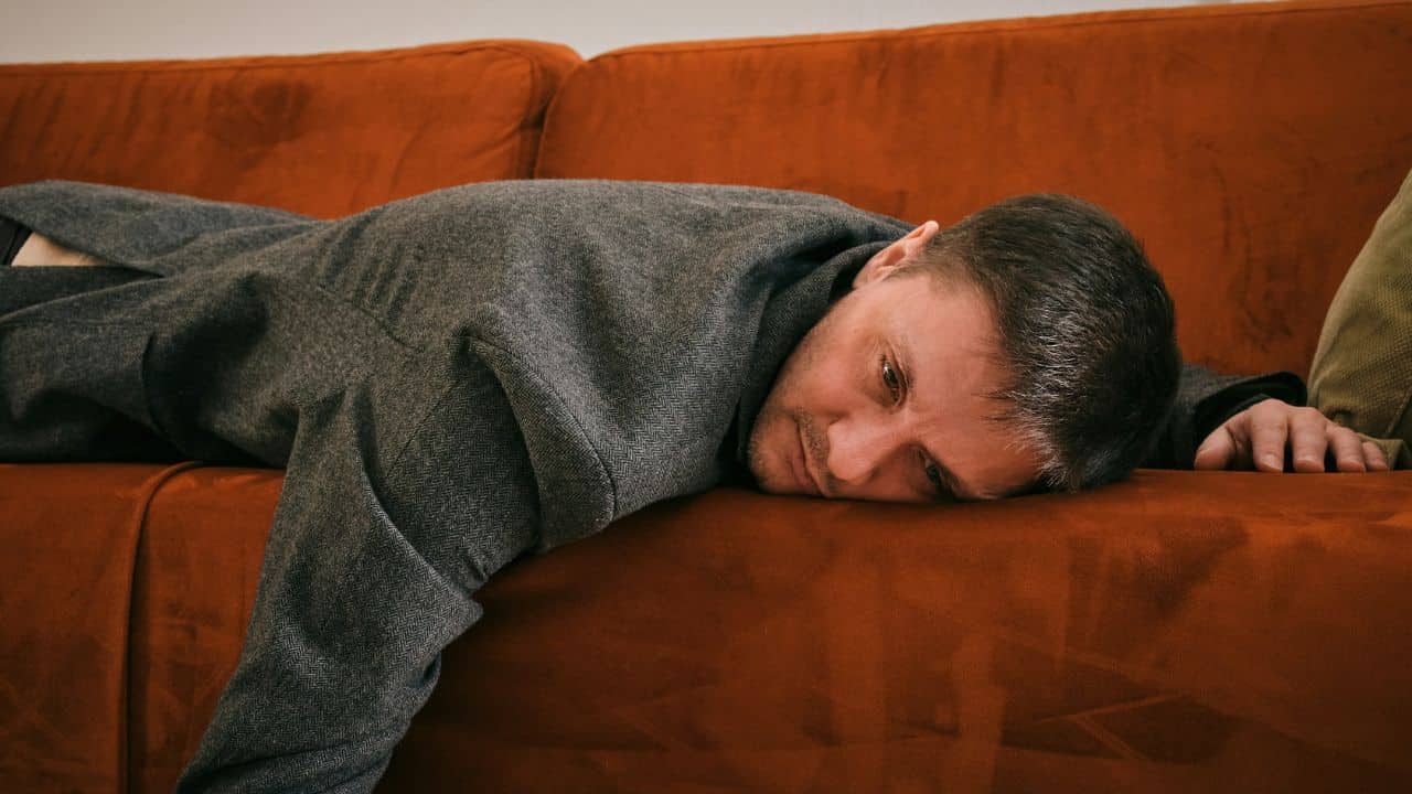 Exhausted man in a gray coat lying face down on an orange velvet sofa.