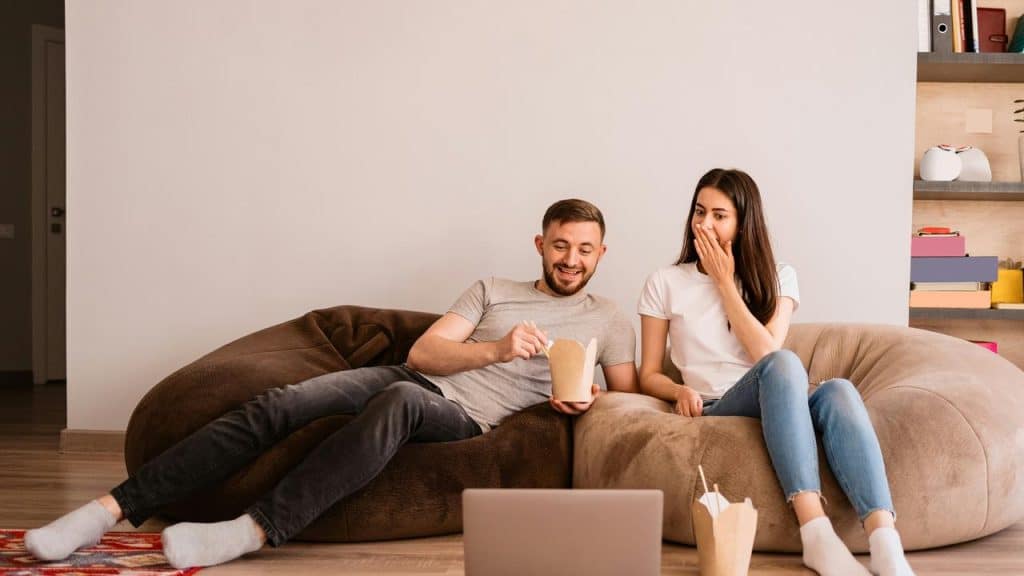 A couple eats takeout and laughs while watching something on a laptop.