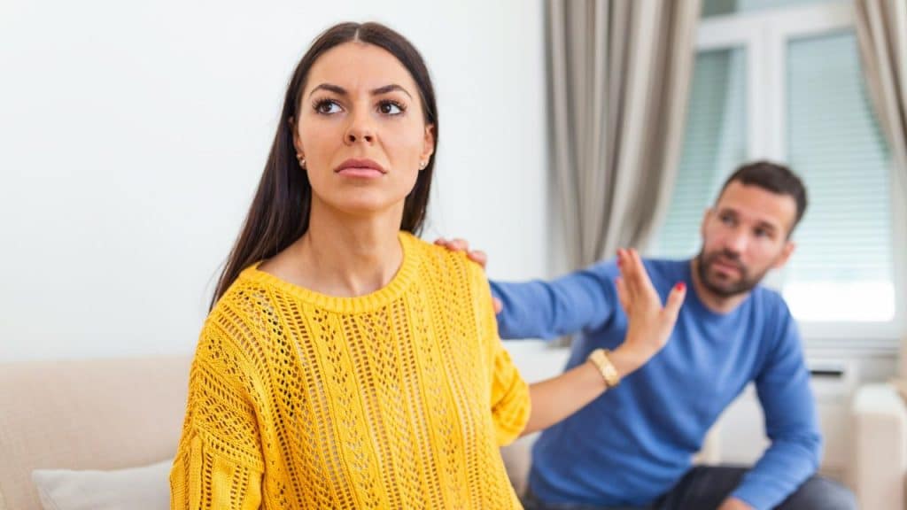 A woman looking away from a man who is reaching out to her shoulder.