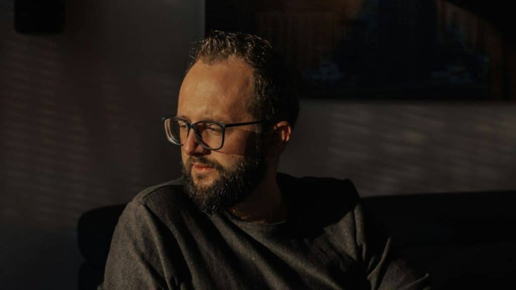 Bearded man in glasses sitting in dim light, looking away from the camera.