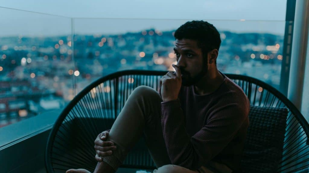 A man sits on a balcony chair at night, looking reflective with a blurred city skyline background.