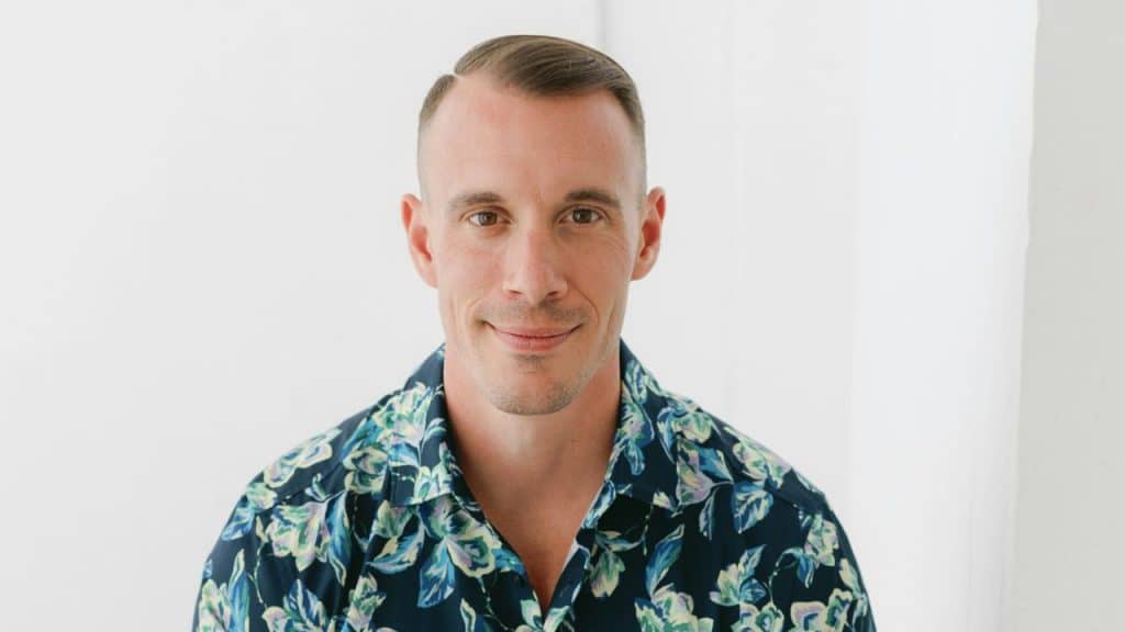 A close-up, headshot of a smiling man with a neat haircut wearing a dark floral shirt.