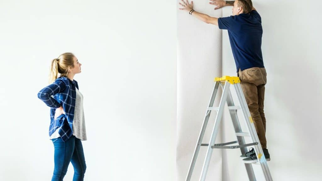 Man on a ladder hanging wallpaper, while a woman sits on the floor watching.