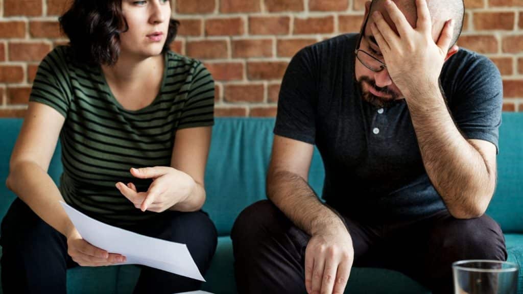 Woman with papers talks to a man with his head in his hand on a couch.