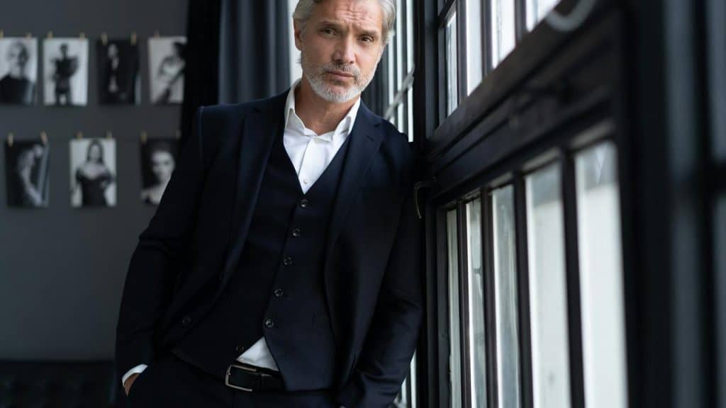 A mature man in a dark suit leaning against a window.