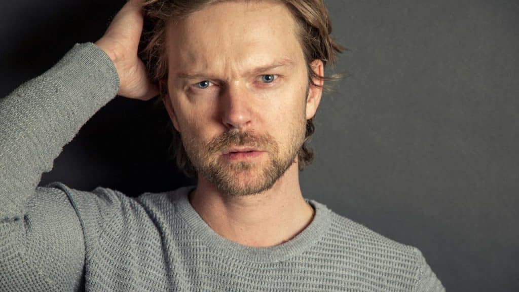 Man in a gray sweater with light hair and a beard, looking intensely at the camera, hand in hair.