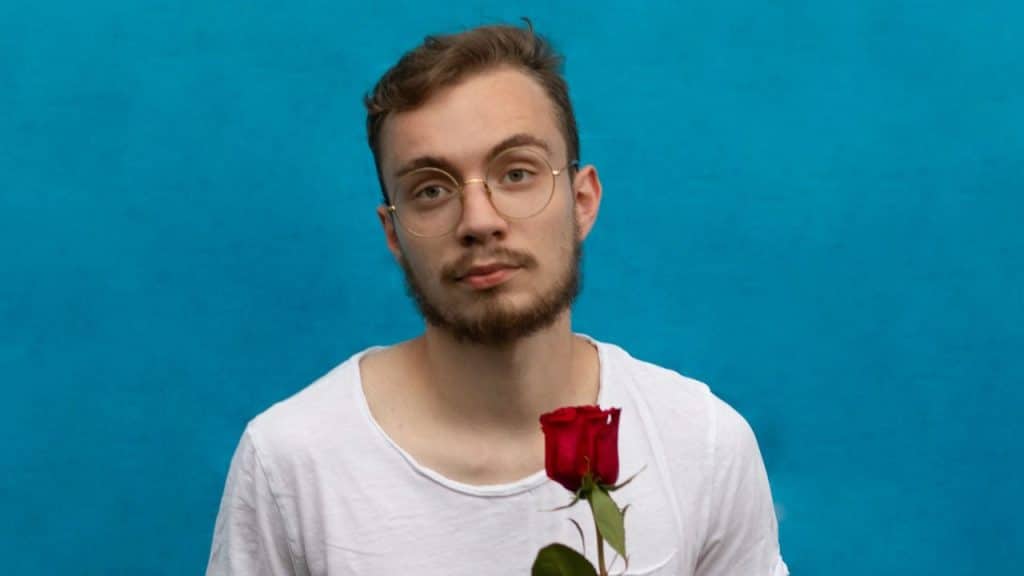 Young man with glasses and a beard holding a single red rose against a blue background.