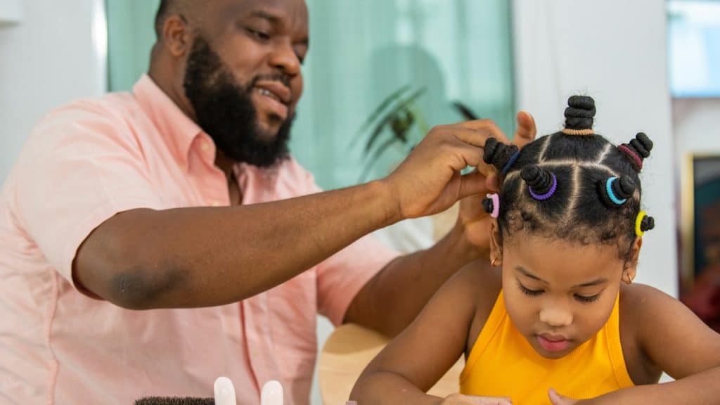 A father tying his little daughter's hair with a colorful hairband in the living room.