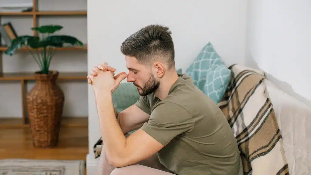 A man sitting on a couch leaning forward with his hands clasped in thought.