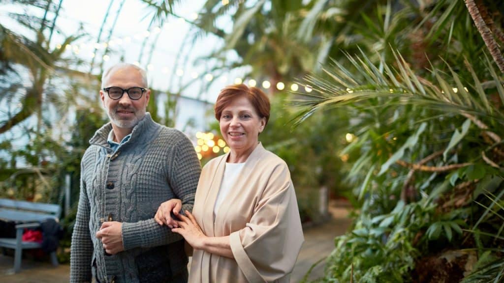 Smiling older couple linking arms and walking through a lush greenhouse.