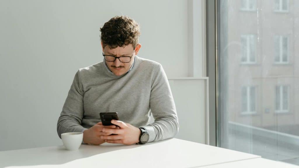 A man wearing glasses sits at a table looking at his phone with a cup nearby.
