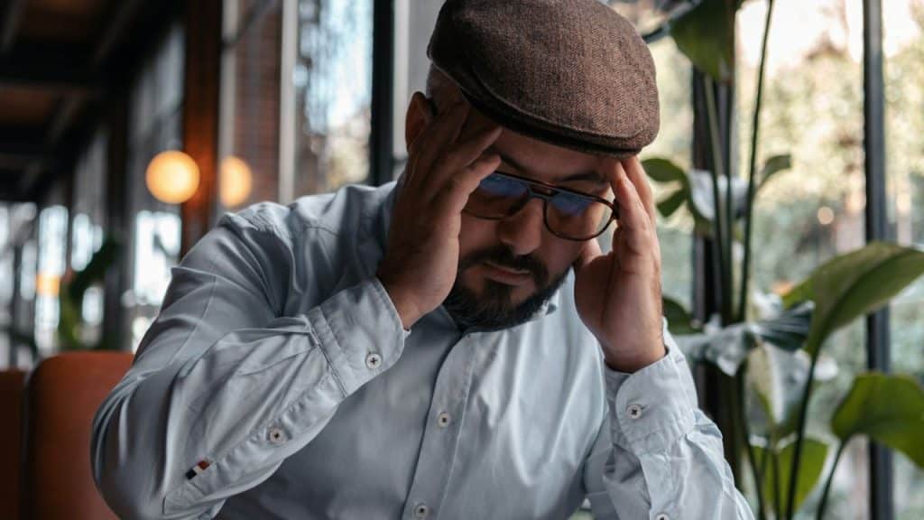 A man in a flat cap, glasses, and a light shirt holds his head in his hands, looking stressed.