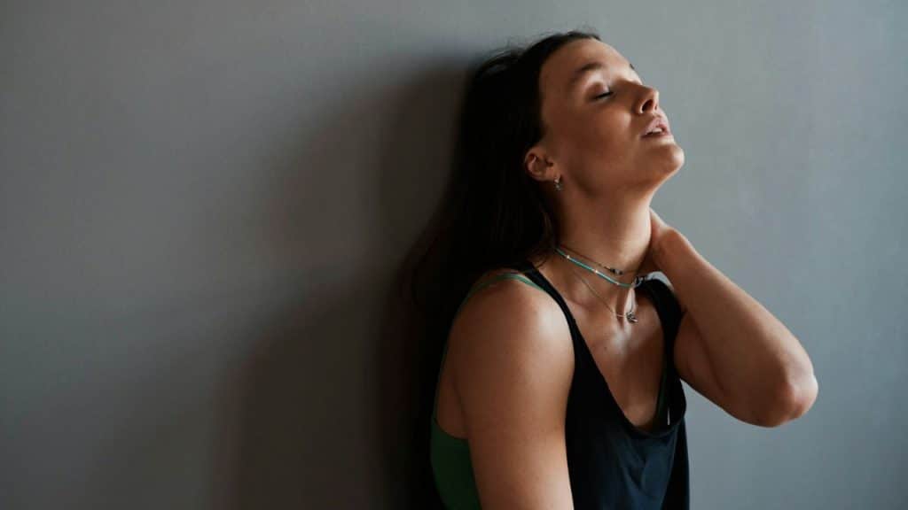A woman leans against a gray wall with her eyes closed and her hand on her neck.