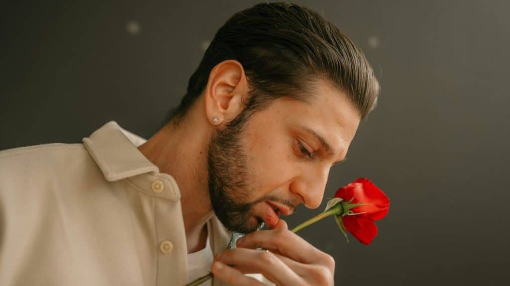 Close-up of a man with a beard smelling a single red rose.