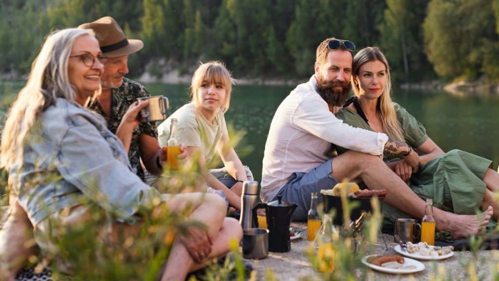 A multi-generational family enjoys a lakeside picnic with food and drinks.