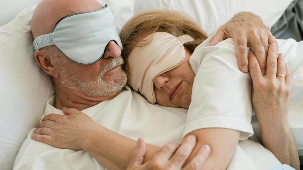 An elderly couple cuddling in bed wearing sleep masks.