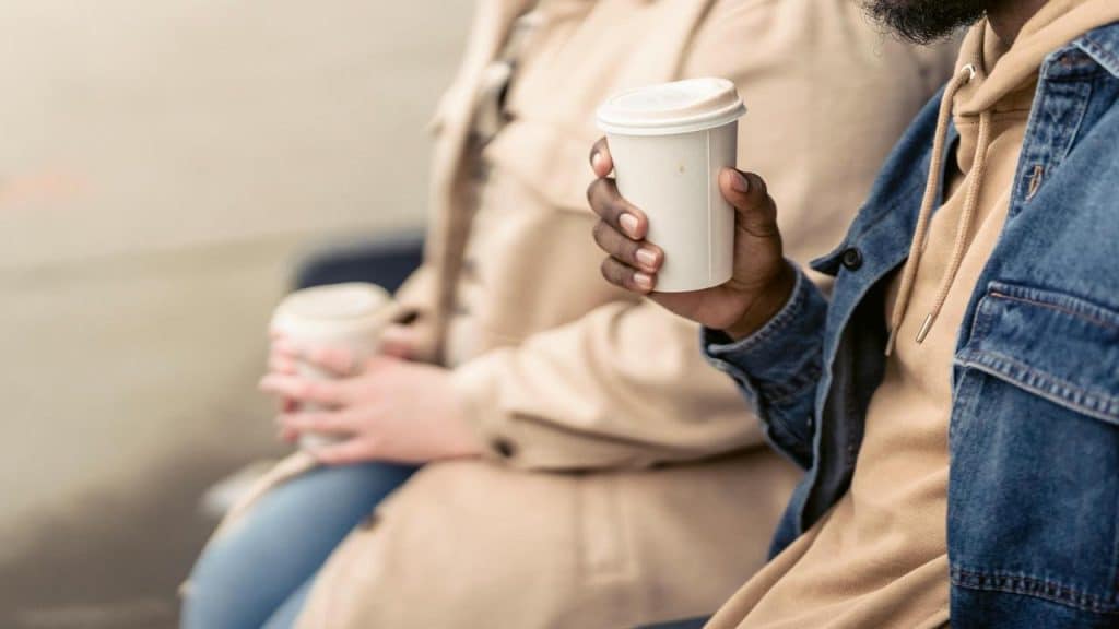 A close-up of two people sitting and holding takeaway coffee cups.