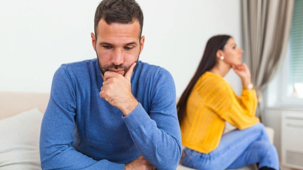A sad man sits in the foreground with a pensive expression, while a woman looks away behind him.
