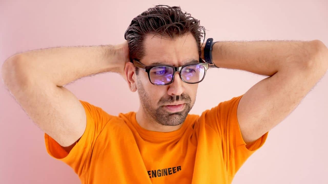 A man in an orange shirt and glasses holds his hands behind his head against a pink background.