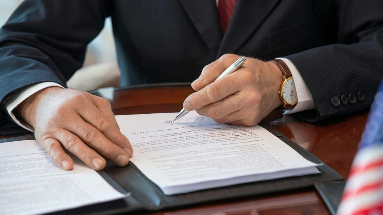 Close-up of a man in a suit signing a document with a white pen.