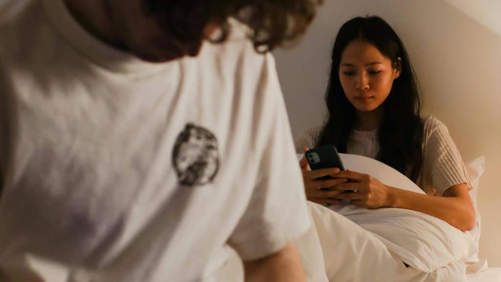A woman sitting in bed looking at her phone while a man sits in the foreground.