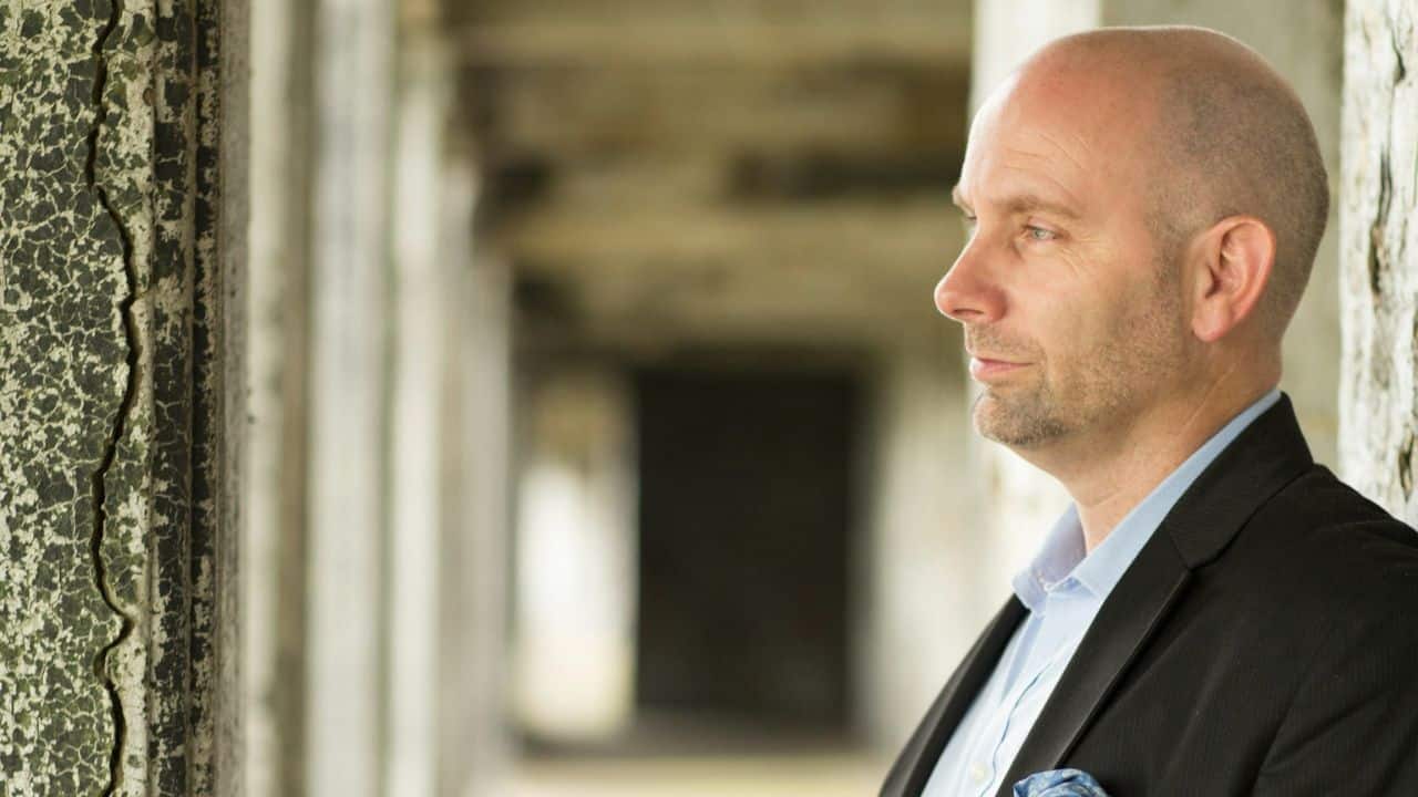 A bald man in a jacket is looking thoughtfully to the right in an old stone structure.