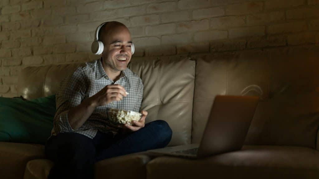 A smiling man wearing headphones is sitting on a couch in the dark, watching a video on a laptop and eating popcorn.
