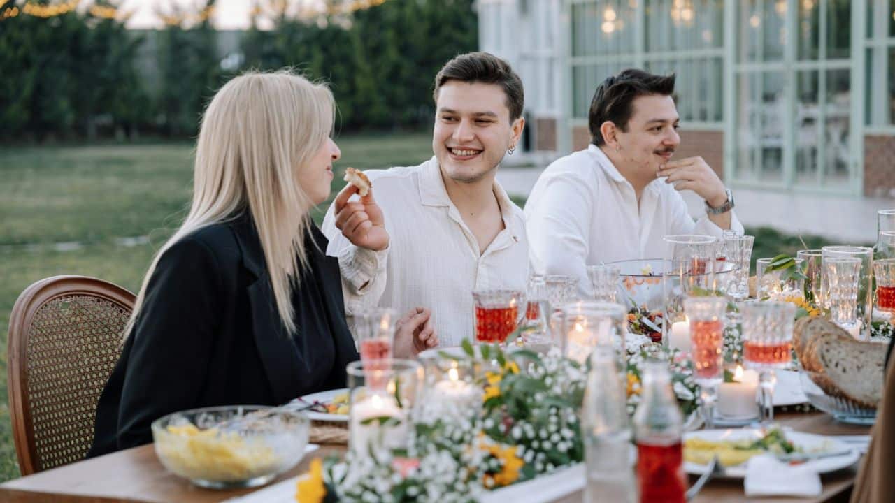 Three people are sitting at an outdoor dining table decorated with flowers and candles.