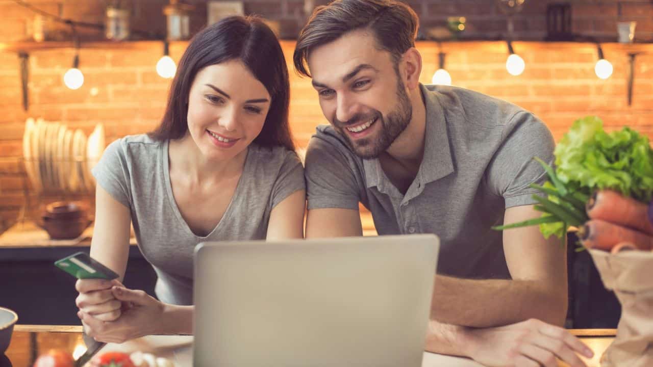 A smiling couple is looking at a laptop in a kitchen, holding a credit card.