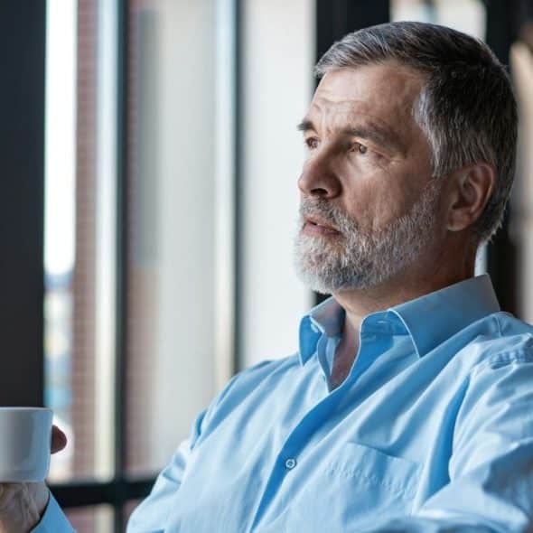 A gray-haired man in a blue shirt holding a white mug and looking out a window.