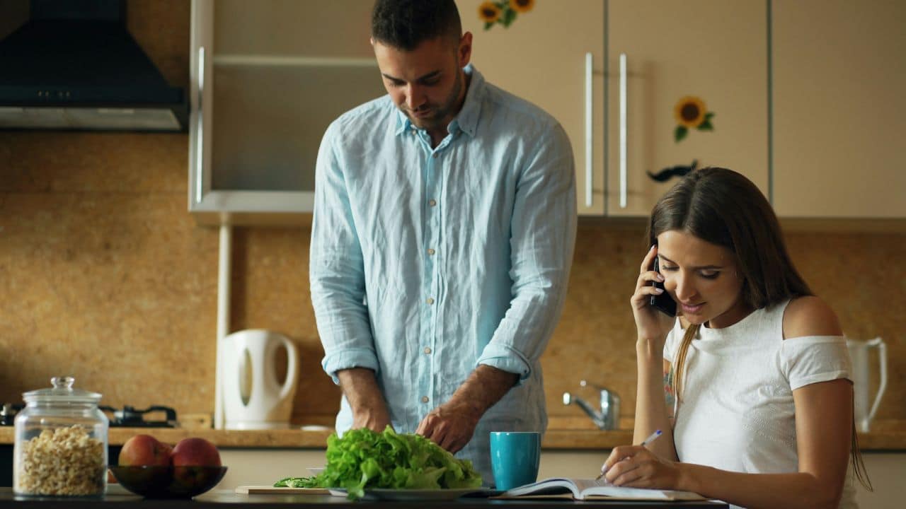 A man chops vegetables while a woman writes and talks on the phone in a kitchen.