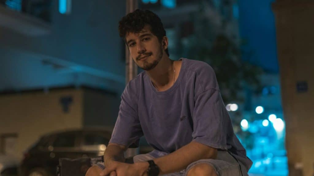 A young man sits outside at night, wearing a purple shirt and looking forward.
