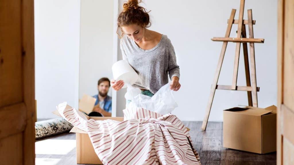 A woman unpacks a box, with a man visible in the background of the room.