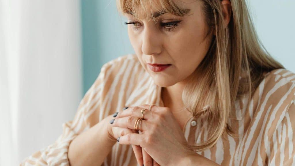 A woman sits with clasped hands, looking down thoughtfully.
