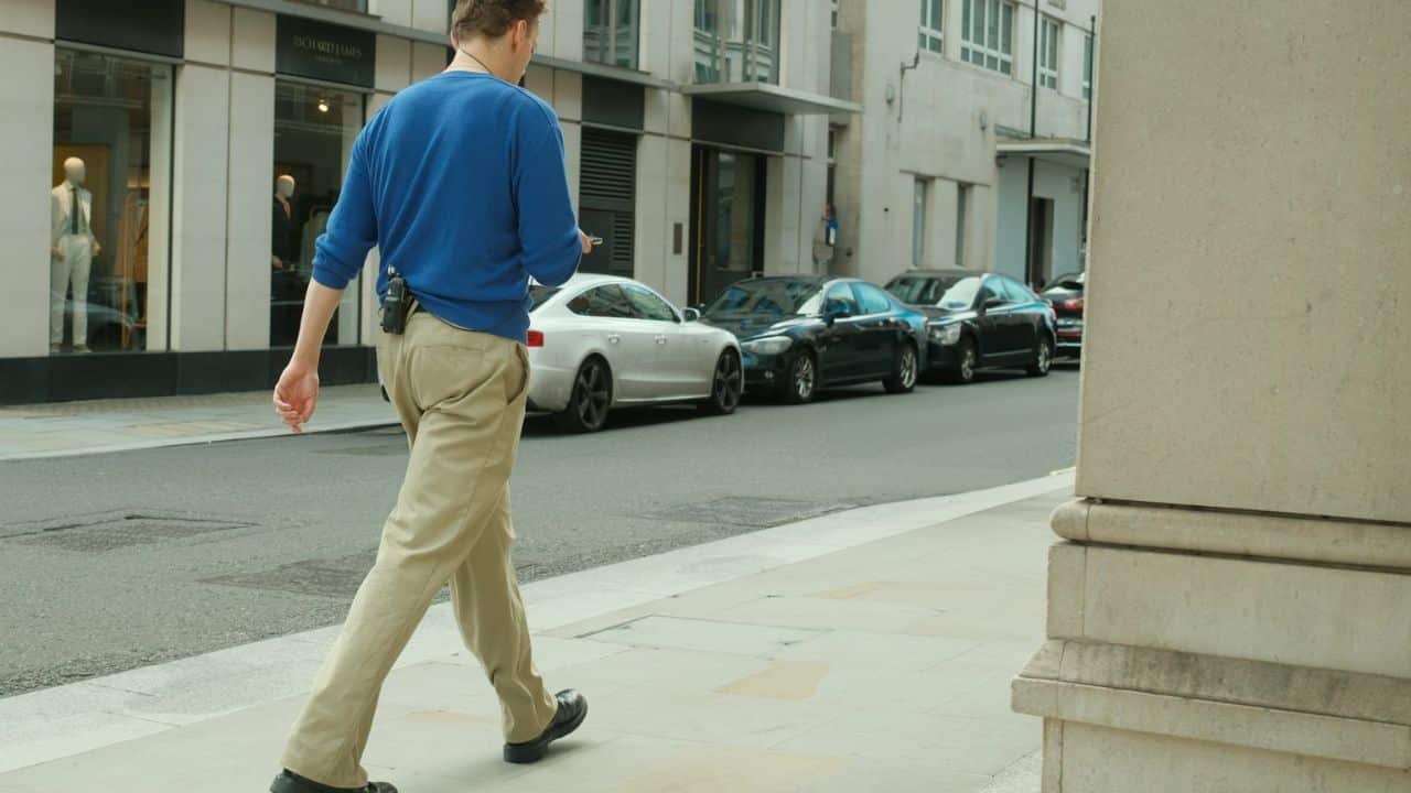 A man in a blue sweater and khaki pants walks along a city sidewalk.