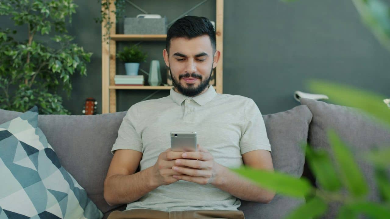 A smiling bearded man sits on a couch looking down at his phone.